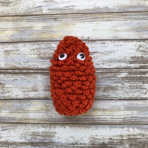 May include: A crocheted orange-brown monster with googly eyes. The monster is made with a textured stitch and has a rounded shape.