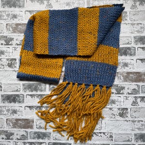 Puede incluir: Bufanda tejida a mano con rayas alternas doradas y azules. La bufanda tiene un borde con flecos de hilo dorado. Hecha de un hilo grueso y texturizado, ideal para el invierno.
