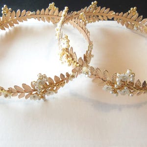 Puede incluir: Dos diademas de metal en tono dorado con un delicado patrón de hojas. Cada diadema está adornada con pequeños detalles florales blancos y racimos de diminutas perlas blancas. Los extremos de las diademas terminan con cintas blancas transparentes.