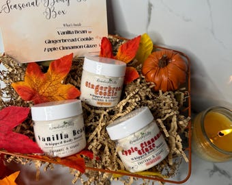 May include: A seasonal gift box with three jars of whipped body butter, a small pumpkin, and fall leaves. The jars are labeled Vanilla Bean, Gingerbread Cookie, and Apple Cinnamon Glaze. The box includes a lit candle.