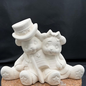 May include: A white ceramic bisque figurine of two teddy bears sitting side-by-side. The bear on the left wears a top hat with stars, and the bear on the right has a bow on its head. They are holding a small flag featuring a star and stripes.
