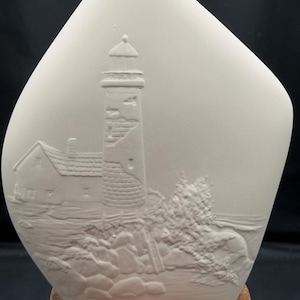 May include: A matte off-white ceramic vase with an asymmetrical shape. The front surface features a raised relief design of a lighthouse, a small house, and rocky terrain with vegetation. The vase rests on a circular, textured cork base.