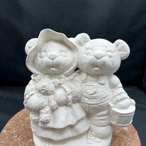 May include: A white ceramic figurine of two teddy bears. One bear wears a bonnet and holds a smaller teddy bear. The other bear wears overalls and carries a small bucket. The figurine is unpainted and ready for customization.