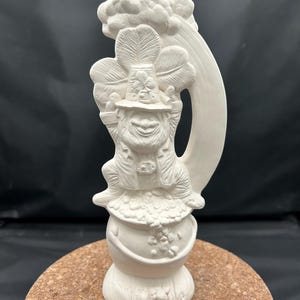 May include: A white ceramic figurine of a leprechaun sitting on a pot of gold. The leprechaun is wearing a hat with a shamrock and holding a large shamrock. A rainbow and cloud are behind the leprechaun.
