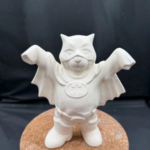 May include: A white ceramic figurine of a bear dressed as Batman, with a mask, cape, and the Batman logo on the chest. The bear is standing on a cork base, arms outstretched. The figurine is unpainted and ready for customization.