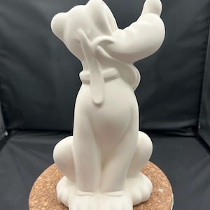 May include: A white ceramic figurine of a dog, seated with its head tilted back and one paw raised. The dog has floppy ears, a collar, and a long tail. The figurine is on a round, cork base.