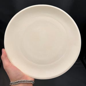 May include: A circular, solid off-white plate with a smooth, matte surface and a simple, clean edge. The plate features a uniform, neutral color throughout its entire surface, presenting a flat and unadorned design for versatile use.
