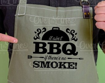 May include: Sage green apron with the text "it ain't BBQ if there's no SMOKE!" in black. The apron has a pocket and adjustable straps. A leather strap detail is visible at the top. The apron is designed for grilling and outdoor cooking.