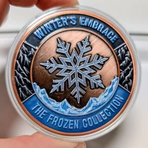 May include: A round, copper-colored coin with a blue and silver snowflake design at the center. The coin has the text "WINTER'S EMBRACE" and "THE FROZEN COLLECTION" in blue. The coin is encased in a clear plastic case.
