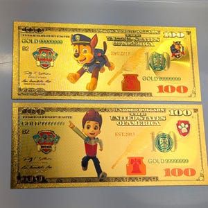 May include: Two rectangular gold-colored novelty bills featuring Paw Patrol characters. The top bill displays Chase, while the bottom bill shows Ryder. Both include the text One Hundred Dollars, The United States of America, Est. 2013, and 100.