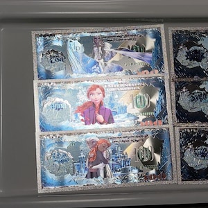May include: Several novelty $100 bills featuring characters from the animated film "Frozen." The bills have a blue and silver color scheme with images of Elsa, Anna, and other characters. The number "100" is prominently displayed.