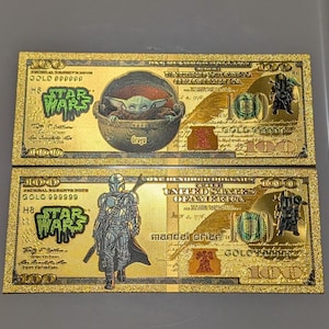 May include: Two gold-colored $100 bills featuring Star Wars characters. One bill shows Grogu in a pod, the other features the Mandalorian. The bills have the Star Wars logo and the text "GOLD" and "100".