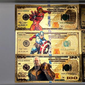 May include: Gold-colored novelty $100 bills featuring Marvel superheroes Iron Man, Captain America, and Thanos. Each bill has the text "GOLD 999999" and "100" printed on it. The bills are displayed in a vertical arrangement.