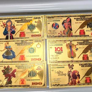 May include: Six gold-colored $100 bills featuring various cartoon characters. Each bill has a unique design with illustrations of characters like Mulan, Alice, and Lady and the Tramp. The bills are decorative and feature the text "GOLD".
