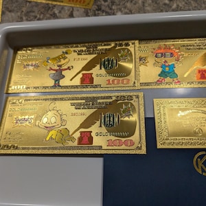 May include: Several gold-colored novelty $100 bills featuring cartoon characters. Each bill has the text "GOLD" and "100" printed on it. The bills are arranged in a gray plastic tray.