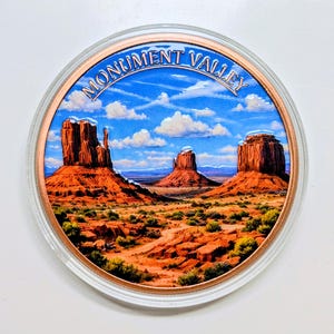May include: A circular commemorative coin featuring a vibrant, detailed illustration of Monument Valley with its iconic red sandstone buttes under a bright blue sky with white clouds. The text MONUMENT VALLEY is printed in blue at the top.