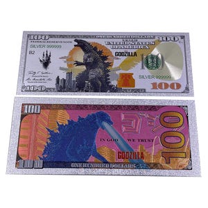 May include: Two decorative silver $100 bills featuring Godzilla. One bill has a Godzilla illustration with a cityscape and the text "GODZILLA". The other bill has a pink background with a blue Godzilla illustration and the text "GODZILLA".