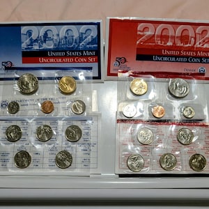 May include: Two 2002 United States Mint Uncirculated Coin Sets. One set is from Philadelphia and the other from Denver. Each set contains various coins in clear plastic packaging, including quarters, dimes, nickels, pennies, and half dollars.
