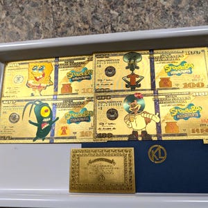 May include: A collection of gold-colored SpongeBob SquarePants novelty $100 bills, featuring characters like SpongeBob, Squidward, and Plankton. The bills are arranged in a tray with a gold plaque and a blue card with a gold emblem.