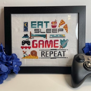 Puede incluir: Un diseño de punto de cruz enmarcado con el texto "Eat Sleep Game Repeat" y varios iconos relacionados con los juegos. El diseño está en un marco negro y está sentado sobre una superficie blanca junto a un mando de Xbox negro.