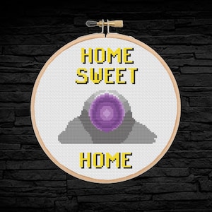 Può includere: Un cerchio da ricamo bianco con un disegno a punto croce che presenta un alieno viola e grigio con il testo "Home Sweet Home" in giallo.
