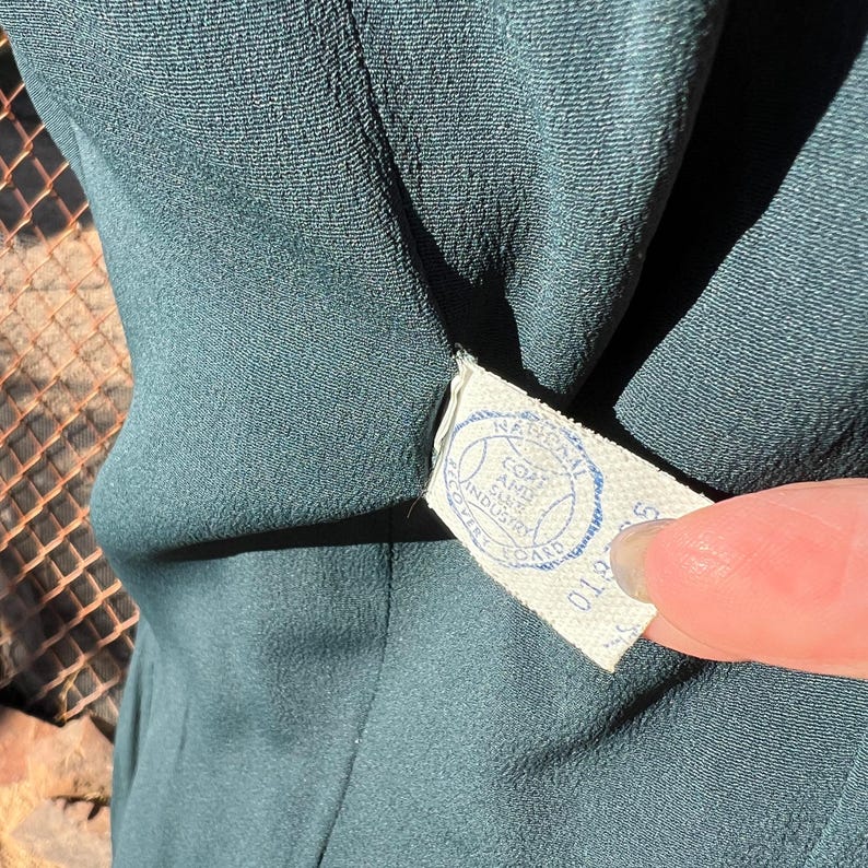 May include: Close-up of a teal-colored garment with a textured surface. A white tag with blue text and a circular logo is attached to the garment. The tag reads "National Coat and Suit Industry Board".