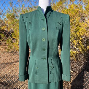 May include: A dark green suit with a mandarin collar, featuring decorative stitching and matching buttons. The jacket has long sleeves with folded cuffs. The suit is displayed on a mannequin.