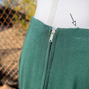 May include: Close-up of a dark green skirt with a visible zipper. The fabric appears to be a solid color. The skirt is on a white form. The background is blurred, with a chain-link fence and foliage visible.