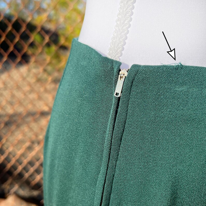 May include: Close-up of a dark green skirt with a visible zipper. The fabric appears to be a solid color. The skirt is on a white form. The background is blurred, with a chain-link fence and foliage visible.