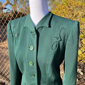 May include: A dark green blazer with a notched collar and decorative button details. The blazer features a series of buttons down the front and decorative cut-out shapes near the buttons. The blazer is displayed on a mannequin.