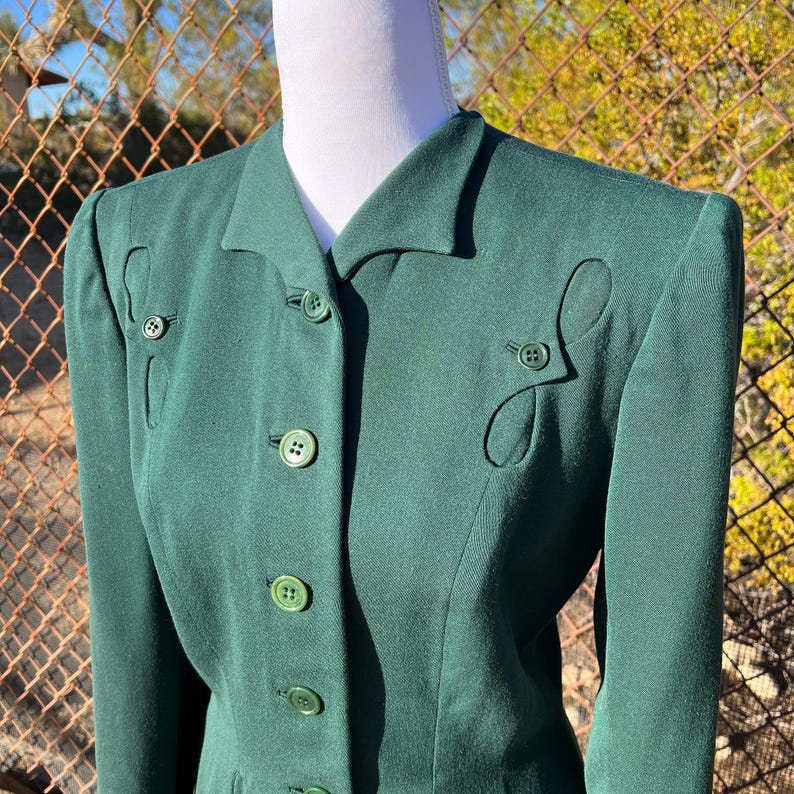 May include: A dark green blazer with a notched collar and decorative button details. The blazer features a series of buttons down the front and decorative cut-out shapes near the buttons. The blazer is displayed on a mannequin.