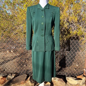 May include: A vintage emerald green skirt suit. The jacket has long sleeves, a notched collar, and decorative stitching. The skirt is knee-length. The suit is displayed on a mannequin against a chain-link fence.