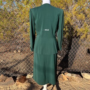 May include: A dark green two-piece suit, consisting of a long-sleeved jacket and a knee-length skirt, displayed on a mannequin. The jacket has a V-neck and the word "HOLE" is printed on the back. The suit is made of a smooth fabric.