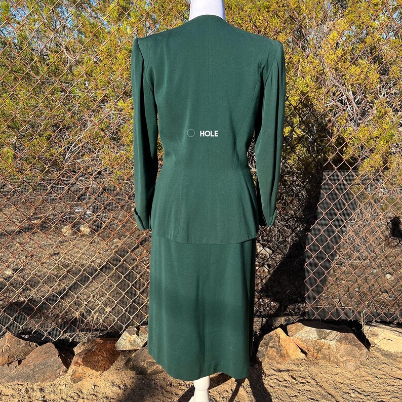 May include: A dark green two-piece suit, consisting of a long-sleeved jacket and a knee-length skirt, displayed on a mannequin. The jacket has a V-neck and the word "HOLE" is printed on the back. The suit is made of a smooth fabric.