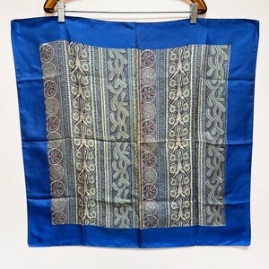 May include: A square silk scarf with a vibrant blue border and a central patterned design. The pattern features vertical stripes with paisley and floral motifs in shades of blue, green, and burgundy. The scarf is displayed on a wooden hanger.