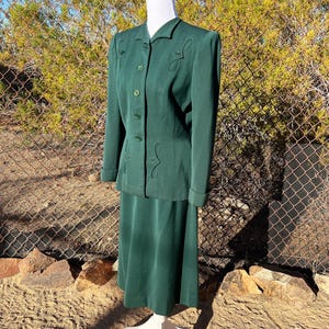 May include: A vintage emerald green skirt suit. The jacket has decorative stitching and button closures. The skirt is a midi length. The suit is displayed on a mannequin. The background includes a chain link fence and natural foliage.
