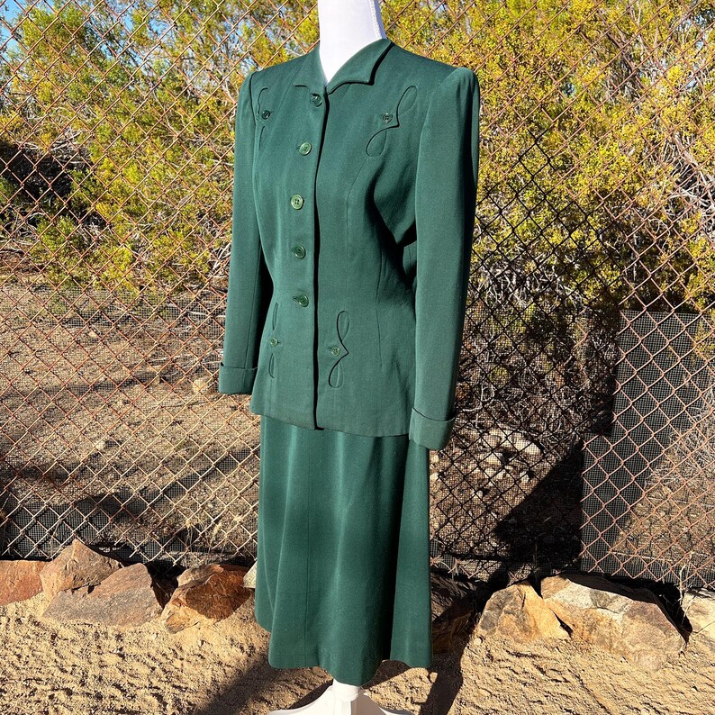 May include: A vintage emerald green skirt suit. The jacket has decorative stitching and button closures. The skirt is a midi length. The suit is displayed on a mannequin. The background includes a chain link fence and natural foliage.