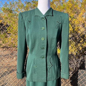 May include: A vintage forest green suit set, featuring a tailored blazer with decorative cutouts and matching skirt. The blazer has a notched collar, button closures, and long sleeves with folded cuffs. The suit is displayed on a mannequin.