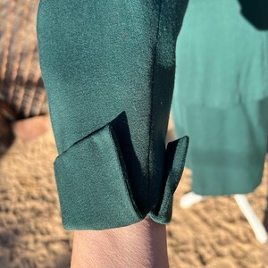 May include: Close-up of a dark green sleeve with a folded cuff. The fabric appears to be a smooth, possibly satin-like material. The cuff is neatly folded, creating a clean, tailored look. The background is blurred, suggesting an outdoor setting.