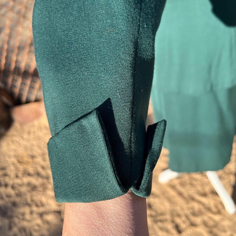 May include: Close-up of a dark green sleeve with a folded cuff. The fabric appears to be a smooth, possibly satin-like material. The cuff is neatly folded, creating a clean, tailored look. The background is blurred, suggesting an outdoor setting.