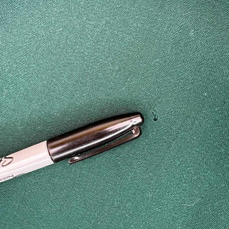 May include: A silver and white Sharpie permanent marker on a green fabric surface. The marker has a silver clip and a black tip. The word "Permanent" is visible on the marker's barrel.