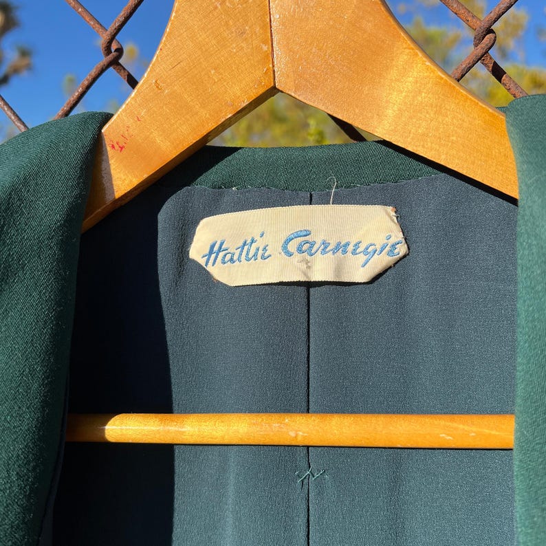 May include: Close-up of a dark green garment hanging on a wooden hanger. The interior lining is a darker shade of green. A label with the text "Hattie Carnegie" in light blue script is visible.