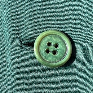 May include: Close-up of a green button with four holes, attached to green fabric. The button is round with a slightly raised edge and a marbled appearance. The fabric has a textured, ribbed pattern.