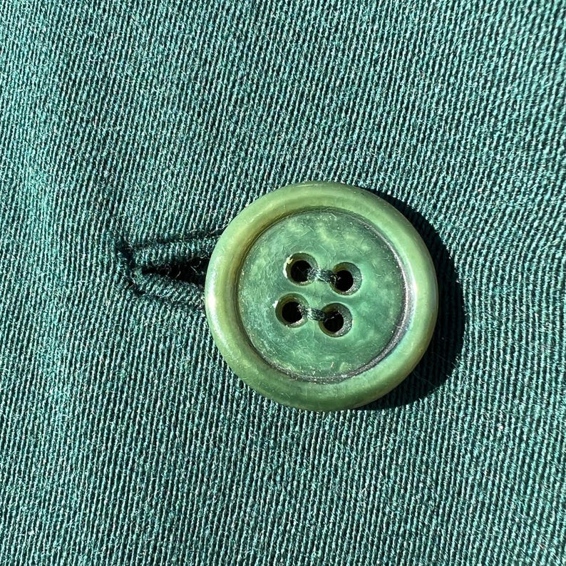 May include: Close-up of a green button with four holes, attached to green fabric. The button is round with a slightly raised edge and a marbled appearance. The fabric has a textured, ribbed pattern.