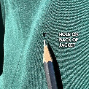 May include: Close-up of a dark green jacket fabric with a small hole. A sharpened pencil points towards the damage. Text on the right reads "HOLE ON BACK OF JACKET."