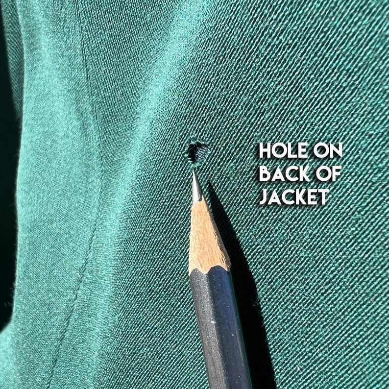 May include: Close-up of a dark green jacket fabric with a small hole. A sharpened pencil points towards the damage. Text on the right reads "HOLE ON BACK OF JACKET."