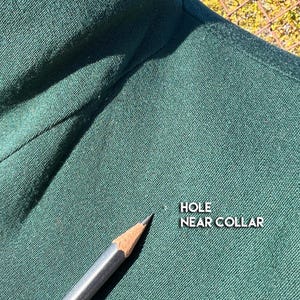 May include: Close-up of dark green fabric with a pencil pointing to a small hole near the collar. The text "HOLE NEAR COLLAR" is visible in white. The fabric appears to be a woven material.