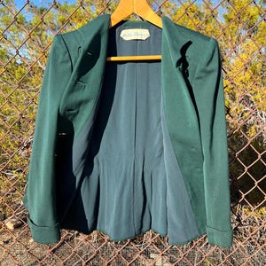 May include: A dark green blazer with a notched lapel and two buttons. The blazer has a classic design with a tailored fit. The interior lining is a darker shade of green. The label inside the blazer reads "Hattie Carnegie".