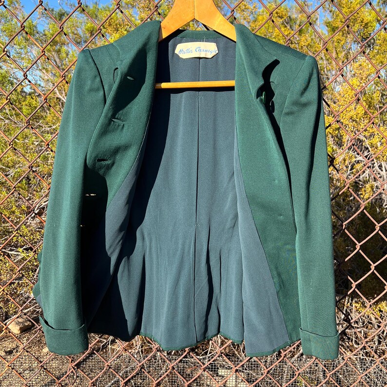 May include: A dark green blazer with a notched lapel and two buttons. The blazer has a classic design with a tailored fit. The interior lining is a darker shade of green. The label inside the blazer reads "Hattie Carnegie".