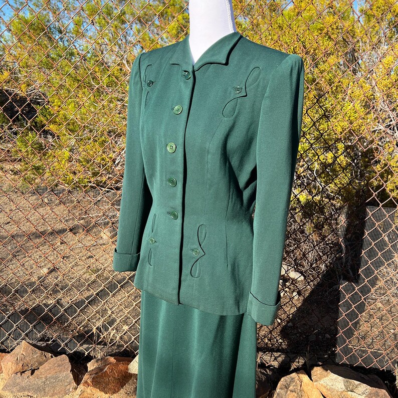 May include: A vintage emerald green skirt suit. The jacket has a notched collar, long sleeves, and decorative stitching. The skirt is a matching solid color. The suit is displayed on a mannequin in front of a chain-link fence.